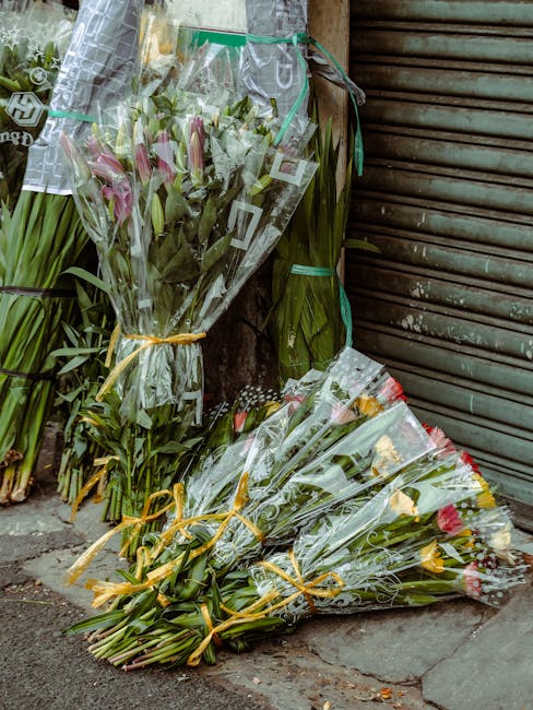 Several bouquets of fresh flowers are placed on the pavement outside a shop, wrapped in clear plastic and tied with yellow ribbon. The arrangements include pink lilies, yellow roses, and various green