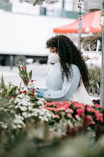 A woman with dark, curly hair wearing a white mask and gloves is selecting fresh flowers at an outdoor flower market. She is dressed in a light blue sweater and appears to be organizing a bouquet. The