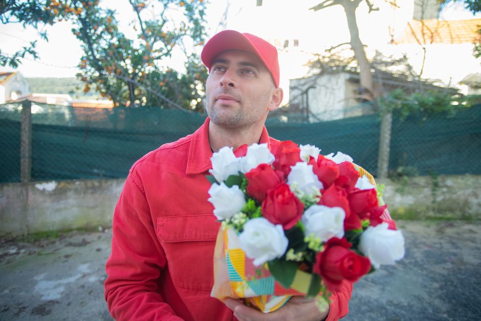 A man dressed in a red jacket and matching red cap is holding a floral bouquet consisting of red and white roses, arranged with green foliage. The bouquet is wrapped in colorful patterned paper, and t