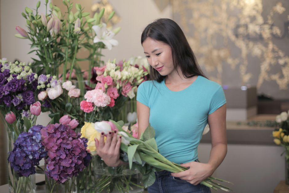 A young woman with long dark hair, wearing a light blue T-shirt, is inspecting a bouquet of fresh flowers inside a flower shop. The arrangement includes pink, purple, yellow, and white flowers such as