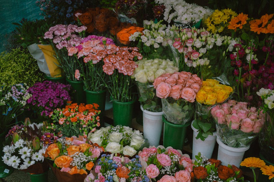 A vibrant display of fresh flowers arranged in green and white buckets and wrapping, featuring pink, white, yellow, orange, and purple blooms. The bouquet includes roses, carnations, daisies, and othe