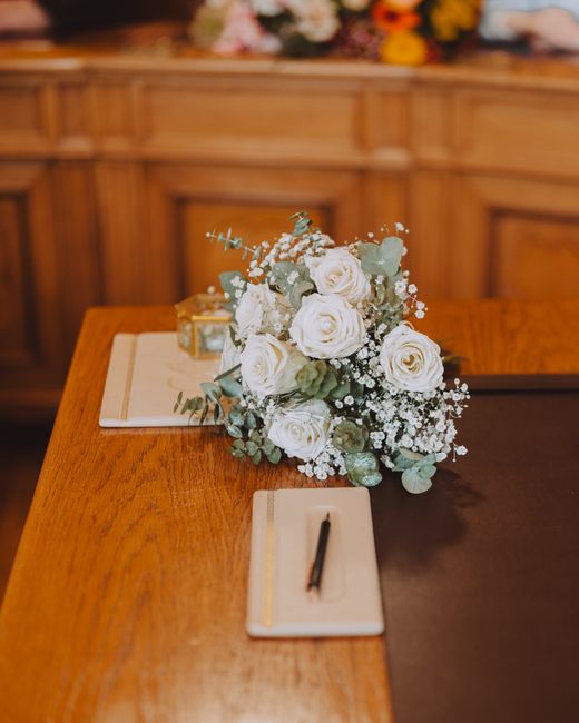 A floral arrangement of white roses, surrounded by delicate sprigs of baby's breath and pale green eucalyptus leaves, artistically placed on a polished wooden table. The bouquet is arranged in a natur
