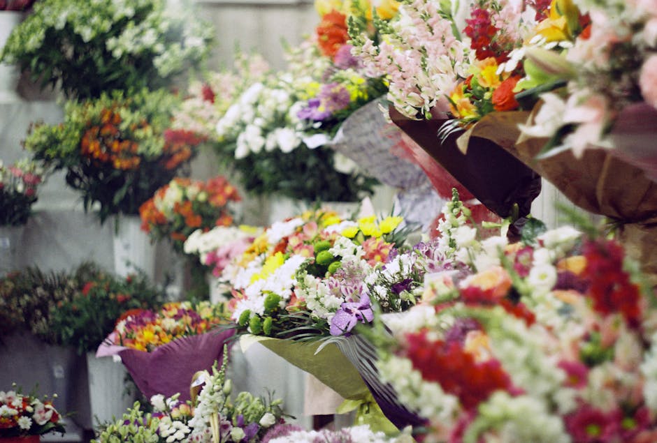 A vibrant display of floral arrangements featuring a variety of fresh flowers including roses, lilies, carnations, and daisies in shades of pink, yellow, purple, orange, and white. The bouquets are wr