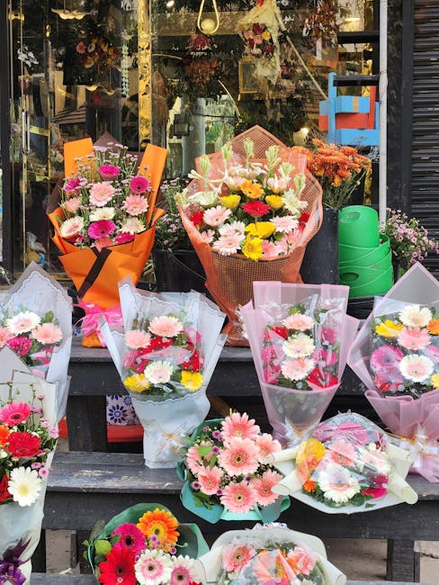 A vibrant display of fresh floral bouquets arranged outside a flower shop on a wooden table and window ledge, featuring a variety of flowers including pink, white, yellow, and red daisies, chrysanthem