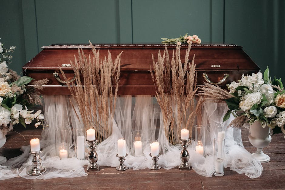 A floral arrangement set up for a funeral or memorial occasion, featuring a closed wooden urn adorned with draped white tulle fabric. In front of the urn are lit white candles in silver and glass hold